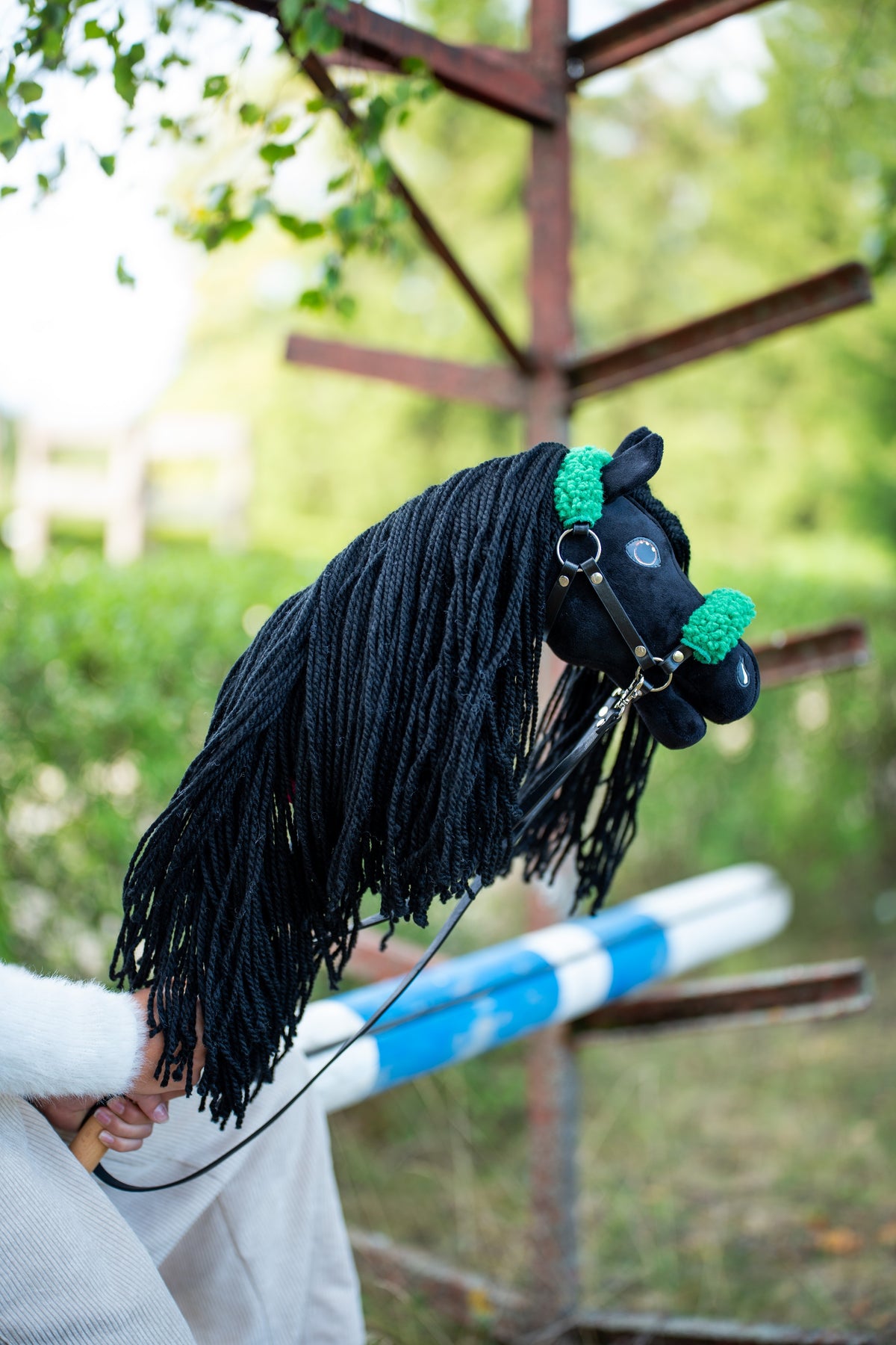 La tête et la longue crinière du Hobby Horse Maximus avec Licol noir de Hobby Horse LarDen sont tenues près d’un obstacle en bois, avec de la verdure derrière, peluche noire et crinière en fil noir, présenté ici avec le licol noir en similicuir à fourrure verte et les rênes noires en similicuir
