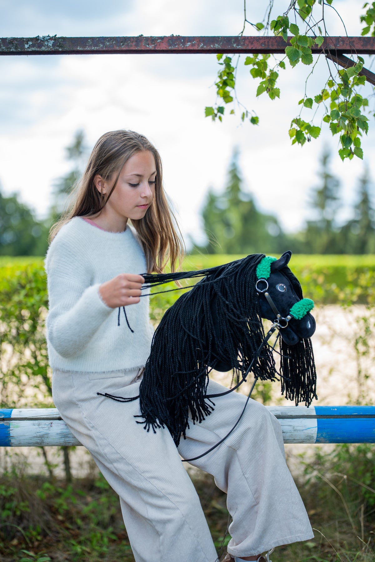 Une fille en pull clair est assise sur une lisse de paddock et rassemble les rênes du Hobby Horse Maximus avec Licol noir de Hobby Horse LarDen, tête en peluche noire et longue crinière en fil noir, présenté ici avec le licol noir en similicuir à fourrure verte et les rênes noires en similicuir