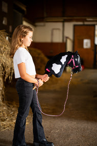 Une fille en haut blanc promène le Hobby Horse Merlin avec Licol noir de LarDen devant des bottes de paille dans une écurie, avec une tête en peluche pie noire et blanche et une crinière en fil noir, présenté ici avec l’ensemble LarDen Exclusive Pink