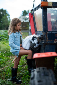 Une fille en veste en jean pose sa botte sur la marche d’un tracteur rouge et noir en tenant le Hobby Horse Nexus avec Licol noir de Hobby Horse LarDen, tête en peluche blanche et crinière en fil blanc, présenté ici avec la bride complète De Luxe à deux frontaux