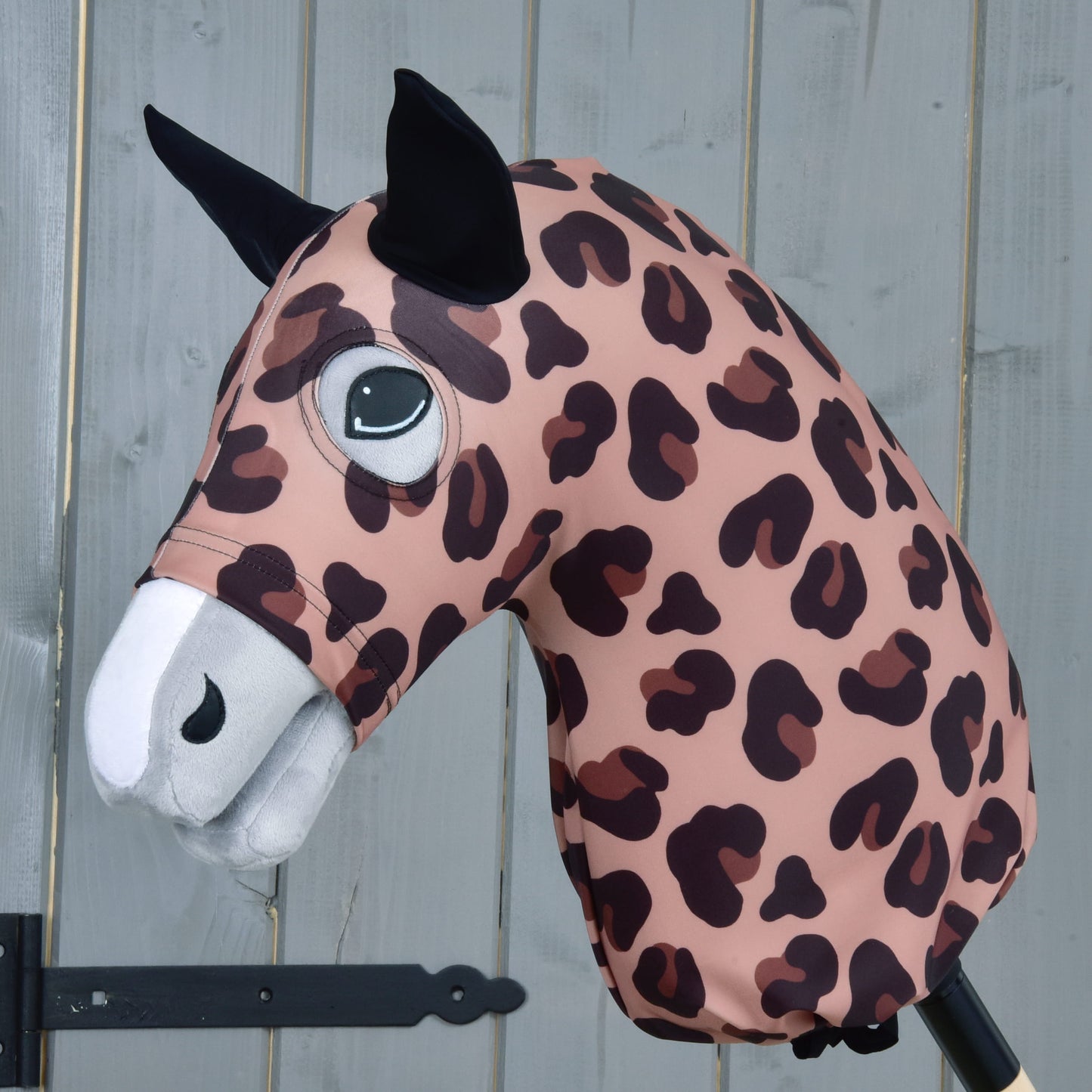 The Hobby Horse LarDen "Hobby Horse protective suit Glamour" features a pink and brown leopard print lycra pattern, black ears, and a white snout, shown on a stick against a gray wooden background.