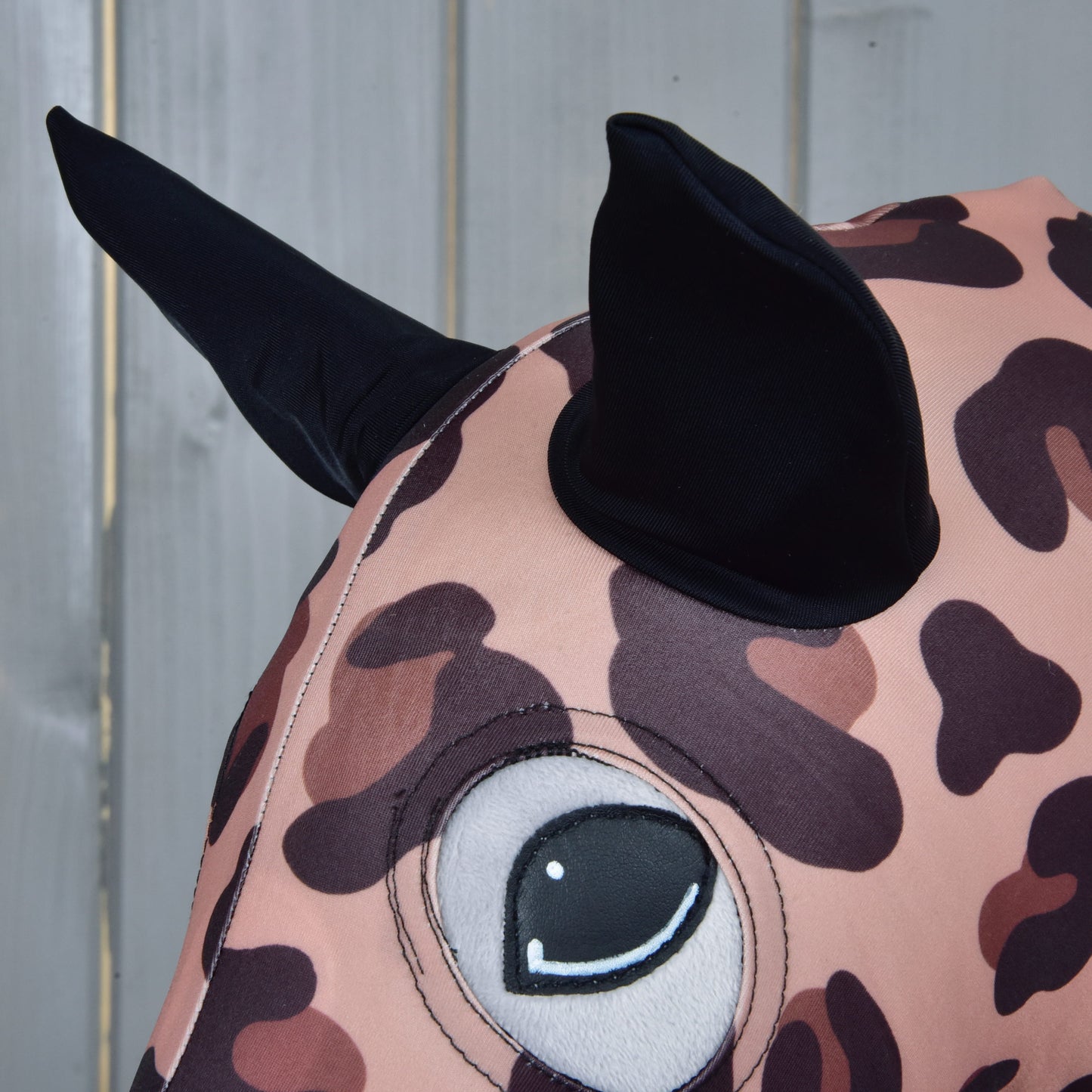 Close-up of the Hobby Horse LarDen "Hobby Horse protective suit Glamour," featuring leopard print fabric, a large stitched eye, and two black pointed ears, resembling a playful hobby horse on a light wooden background.