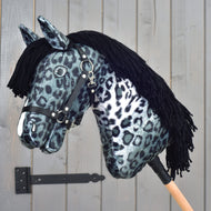 Hobby Horse Picasso avec licol noir de Hobby Horse LarDen, présenté comme un étalon saisissant, avec un tissu noir et blanc à motif léopard rappelant la robe tachetée d’un Appaloosa, une crinière en fil noir et un licol noir, monté sur un bâton en bois devant une porte en bois grise.