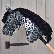 Hobby Horse Picasso avec licol noir de Hobby Horse LarDen, avec tête en peluche à robe tachetée grise et noire inspirée de l’Appaloosa, licol noir et crinière en fil noir, monté sur un bâton en bois et présenté devant un mur en bois gris.