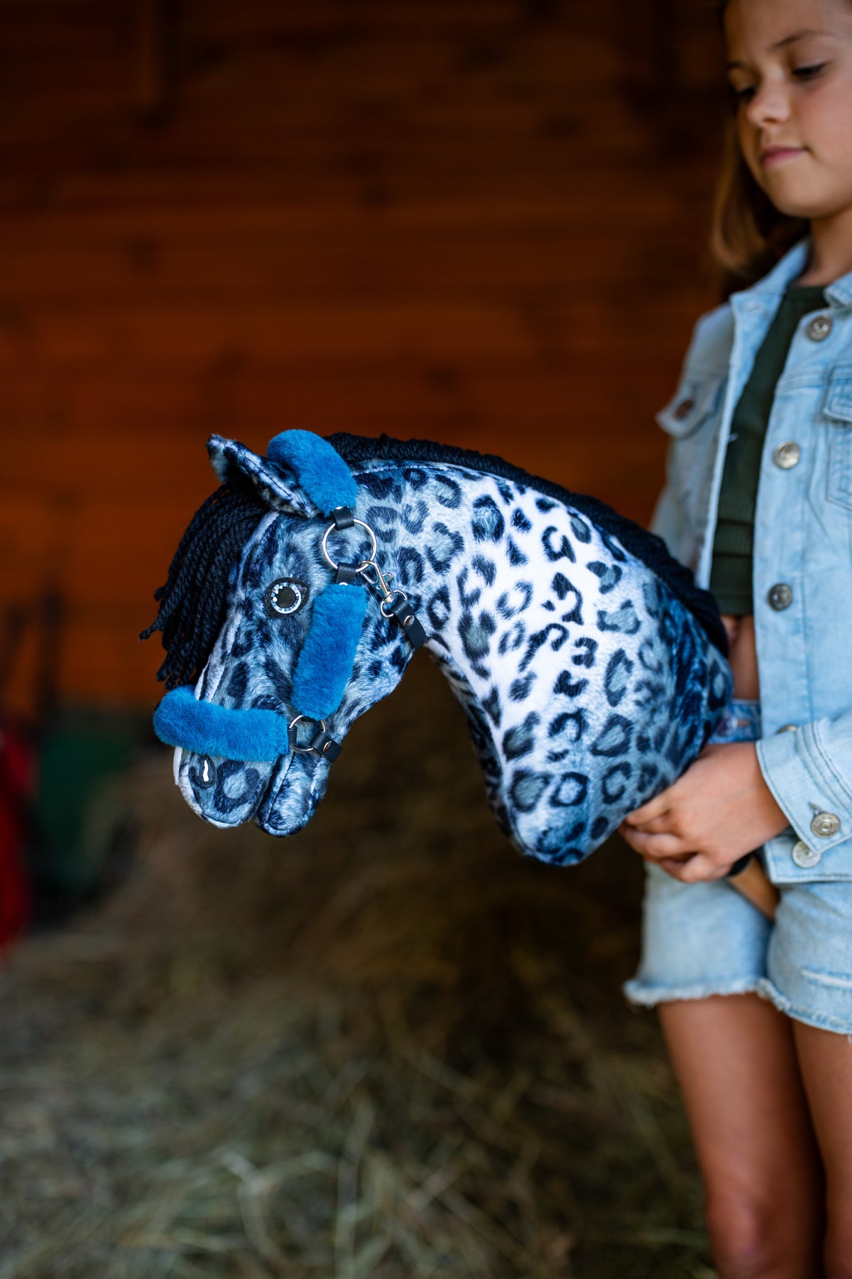 Une fille en veste en jean tient le Hobby Horse Picasso avec Licol noir de Hobby Horse LarDen contre un mur de grange rouge en bois, tête en peluche à imprimé léopard bleu et blanc et crinière en fil noir, présenté ici avec le licol Teddy Petrol doublé de fourrure