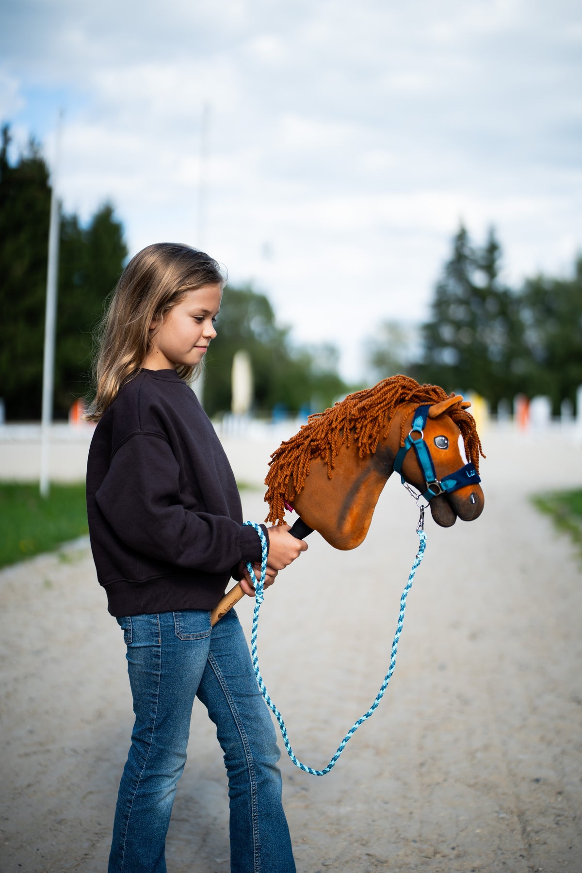 Une fille en sweat sombre promène le Hobby Horse Pippa avec Licol noir de Hobby Horse LarDen sur un chemin sablonneux près de la carrière, tête en peluche alezan et crinière en fil roux, présenté ici avec le licol LarDen Blue doublé de fourrure et la longe Bloom Blue