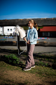 Une fille en veste en jean claire et pantalon mauve se tient devant une clôture de paddock en bois avec le Hobby Horse Pixie avec Licol noir de Hobby Horse LarDen, tête en peluche beige à crinière en fil blanc, présenté avec le Licol gris avec fourrure rose et la Longe rayée Tizian