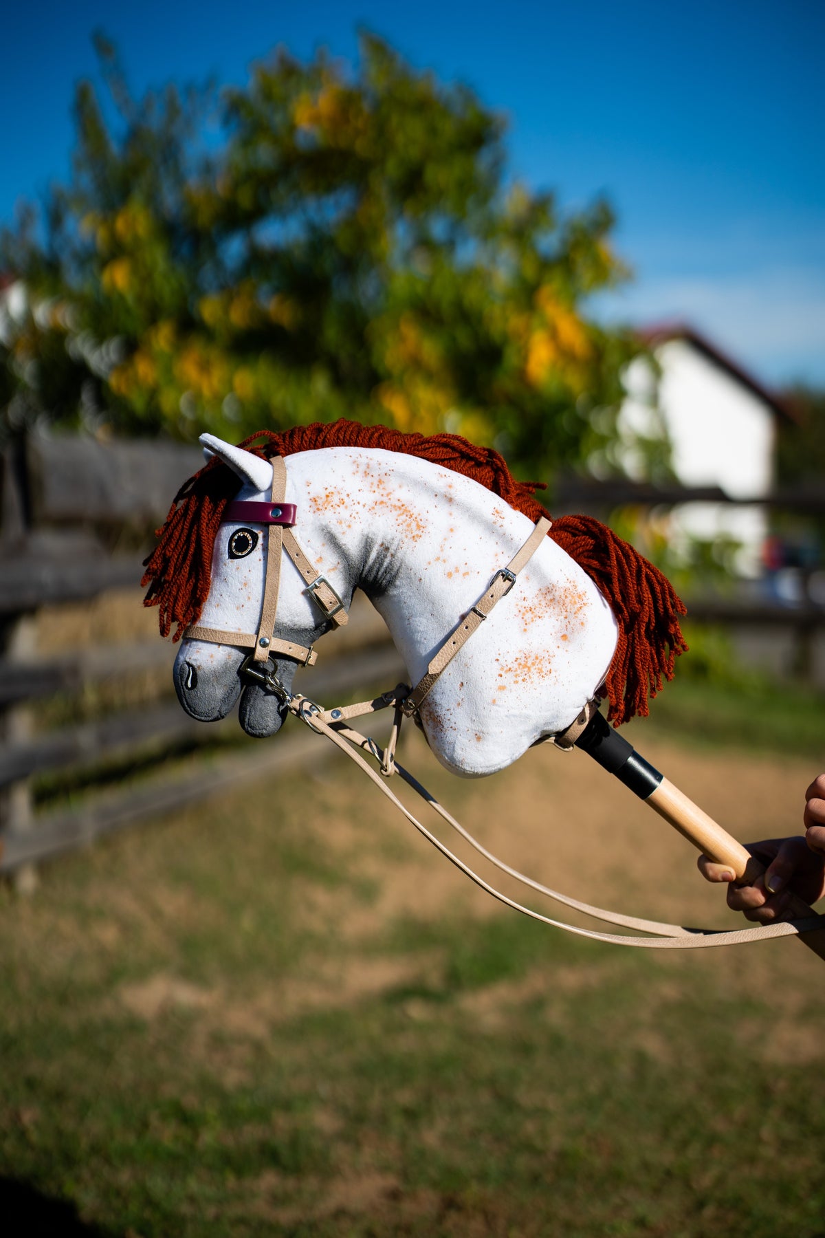 Hobby Horse blanc moucheté à crinière en laine brun-rouge avec bride et rênes beiges et frontal bordeaux près d’une clôture de paddock