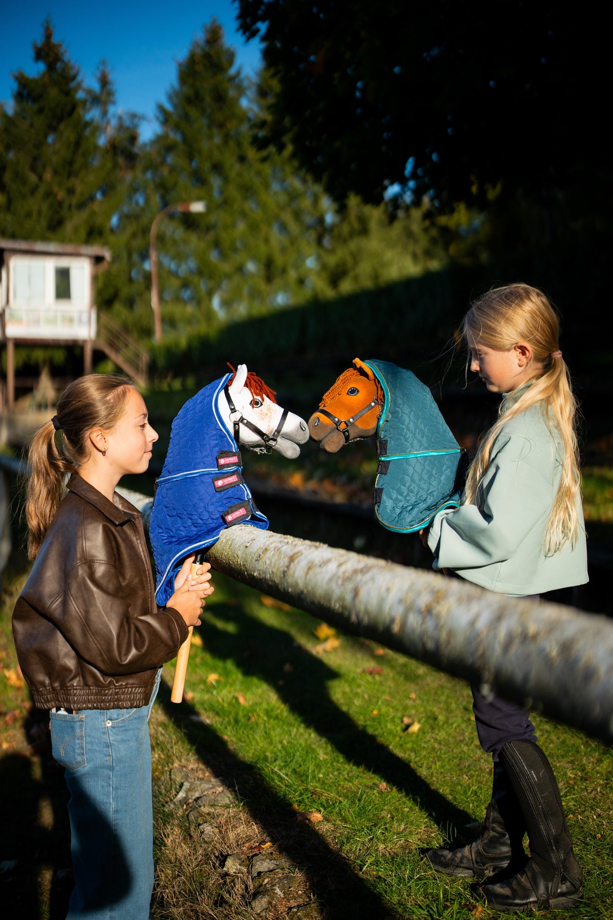 Deux filles près d’une barrière en bois tiennent face à face au soleil couchant le Hobby Horse Rufus avec Licol noir dans la Couverture avec couvre-cou amovible Bluebell et le Hobby Horse Pippa avec Licol noir dans la Couverture avec couvre-cou amovible Lagoon, tous deux de Hobby Horse LarDen