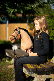 Une fille en pull noir est assise sur un banc de parc au soleil du soir, tenant le Hobby Horse Samantha avec Licol noir de Hobby Horse LarDen, tête en peluche sable et crinière en fil noir, présenté ici avec la bride noire et ses rênes à lanières