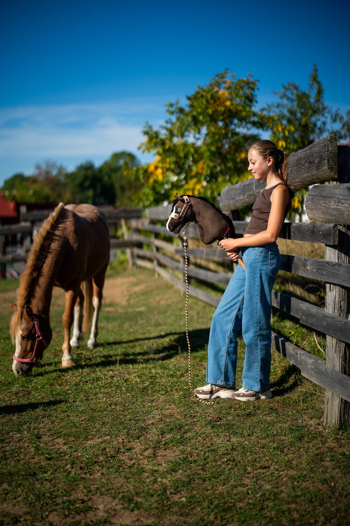 Fille en jean à la clôture du paddock tenant le Hobby Horse Sisi en longe pendant qu’un poney alezan broute à côté