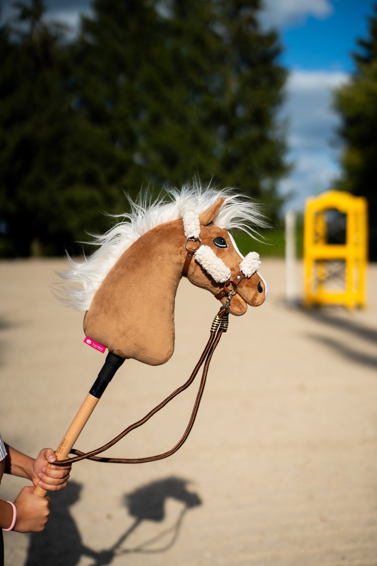 La tête et la crinière soufflée par le vent du Hobby Horse Star avec licol noir de Hobby Horse LarDen sont tenues sur une carrière en sable, peluche palomino, crinière blanche en fil et muserolle en fourrure blanche, présenté ici avec le licol marron à fourrure intégrale et les rênes Vivaldi