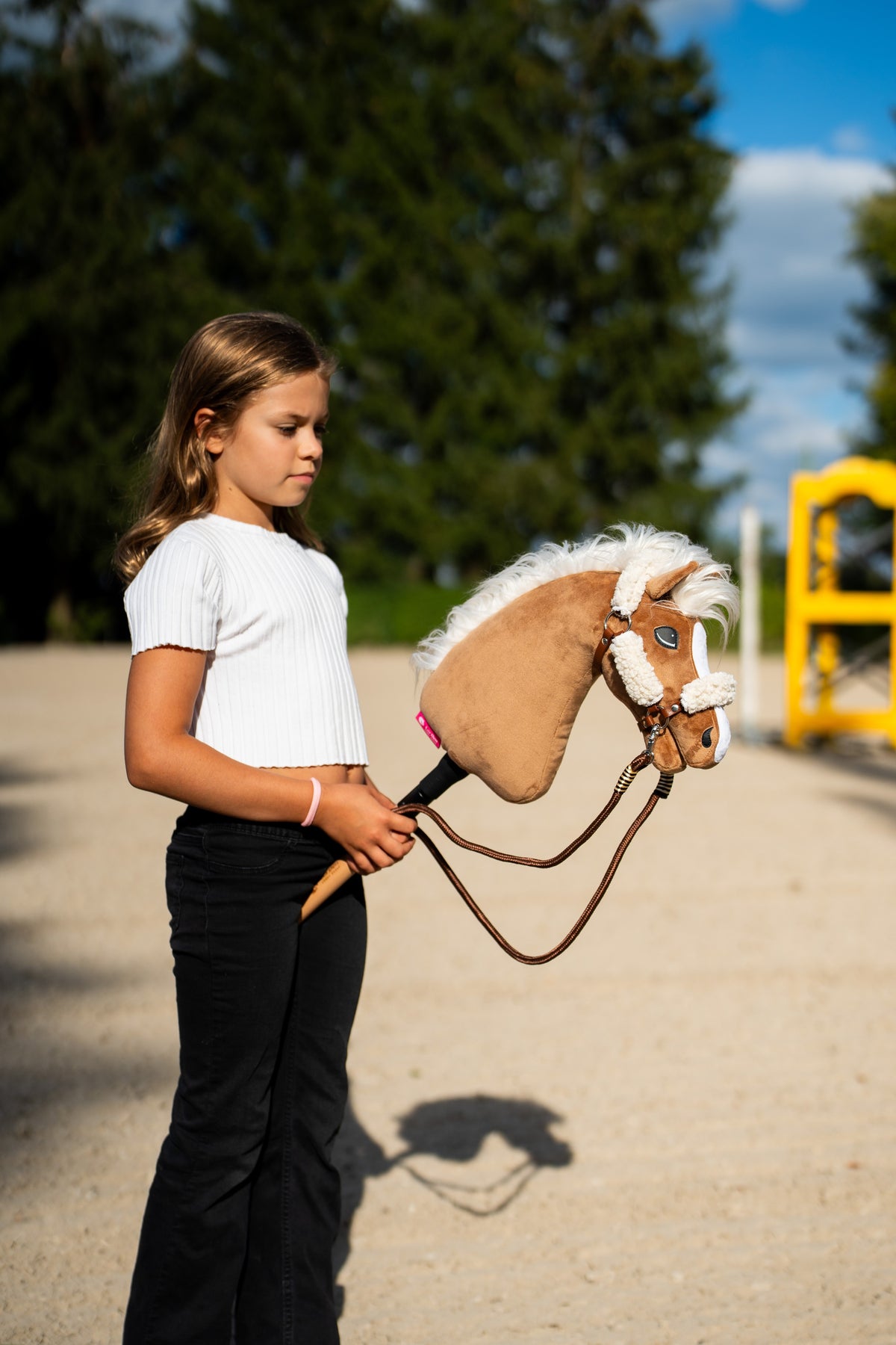 Une fille en haut blanc promène le Hobby Horse Star avec licol noir de Hobby Horse LarDen sur une carrière en sable, avec un chandelier d’obstacle jaune derrière, tête en peluche palomino et crinière blanche en fil flottante, présenté ici avec le licol marron à fourrure intégrale et les rênes Vivaldi