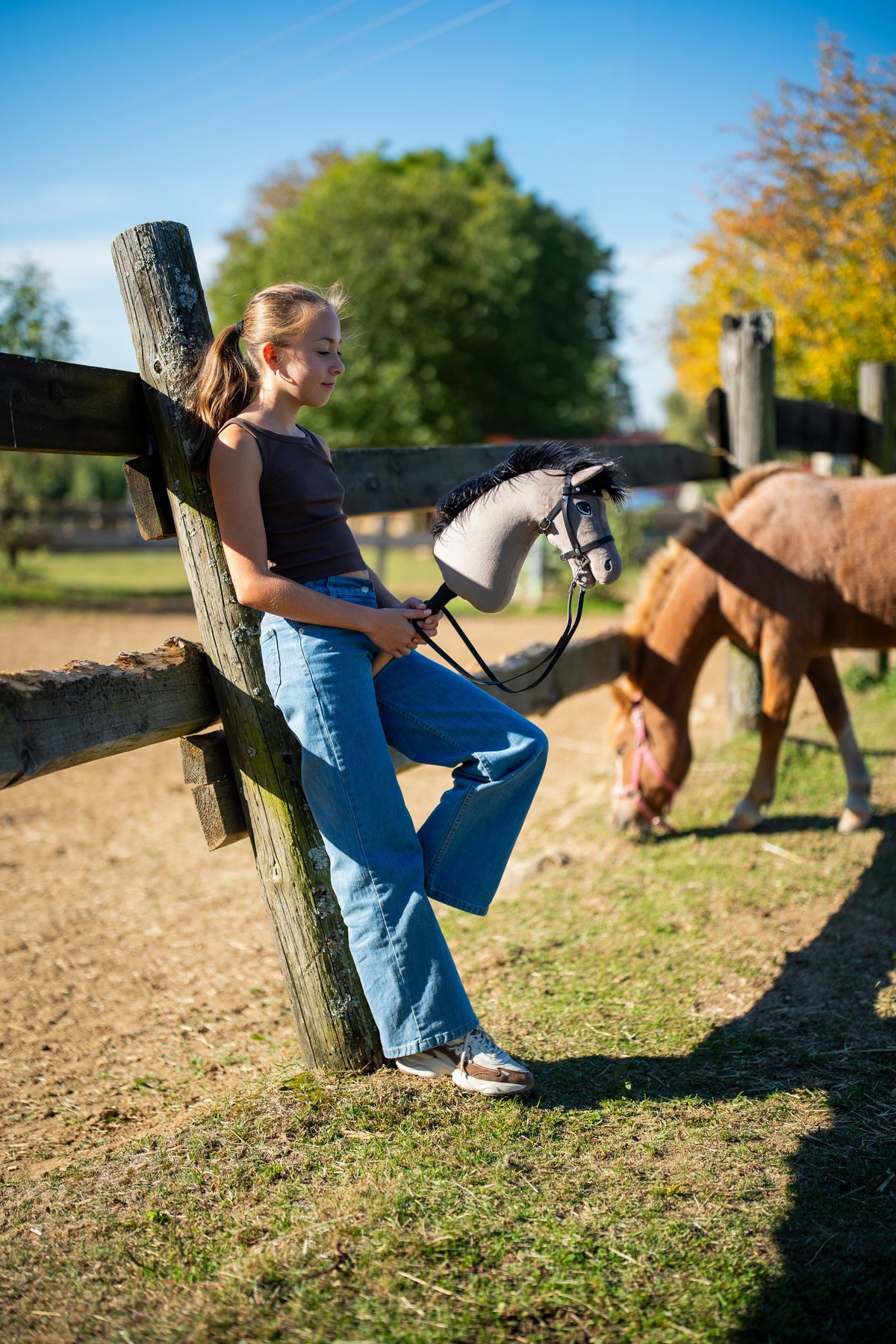 Fille appuyée sur une clôture de paddock au soleil d’automne avec le Hobby Horse Thor en bride noire, un poney alezan broutant derrière