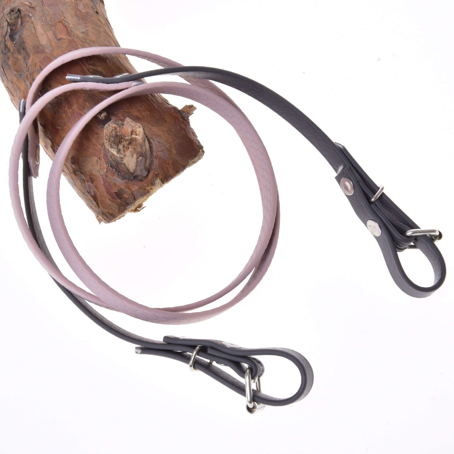 A light brown wooden branch with two Hobby Horse LarDen Reins rubberized Dark Grey/Pink looped around it, resembling leather dog leashes, against a white background.